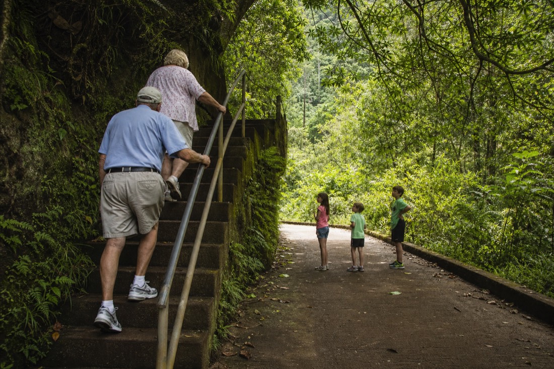 Grandparents using the handrail on a shaded stair flight while children wait on the paved trail below