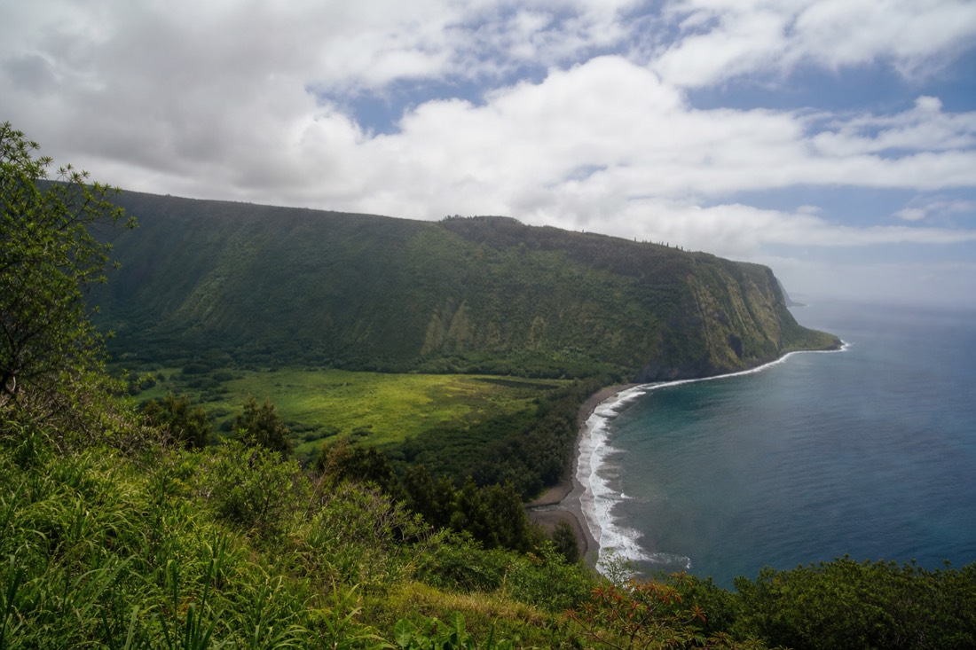 Waipiʻo Valley Lookout on the Hamakua Coast with green cliffs falling to a black sand shoreline under scattered cloud