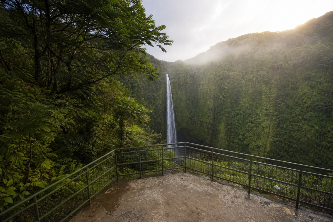 Empty railed overlook platform facing a tall narrow waterfall dropping into a deep green gorge, early morning sun catching the far ridge above the mist