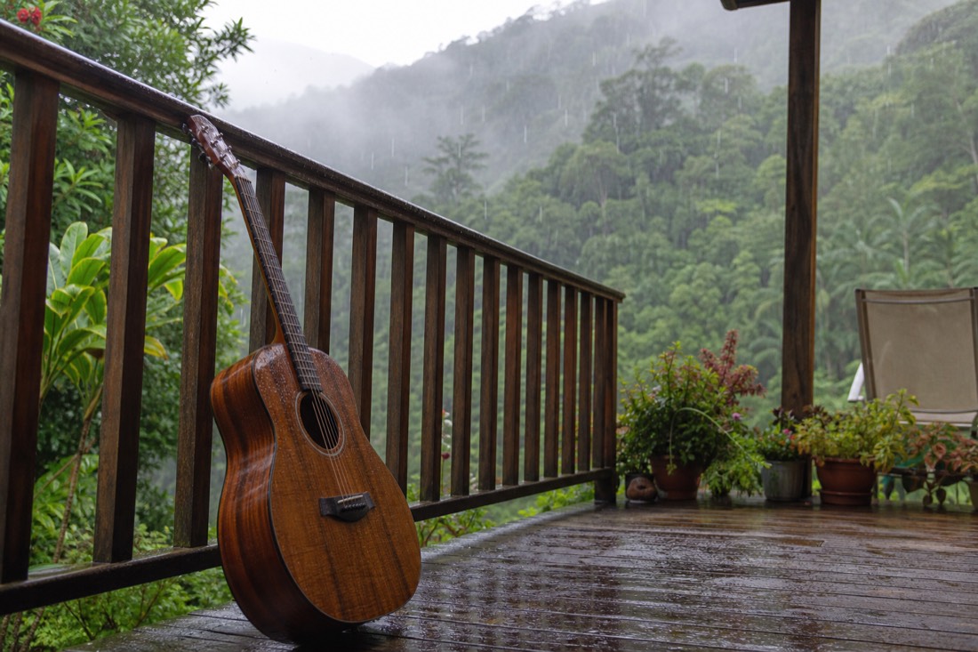 A koa-bodied acoustic guitar resting against the rail of a weathered wooden lanai with heavy rain falling on green hillside beyond