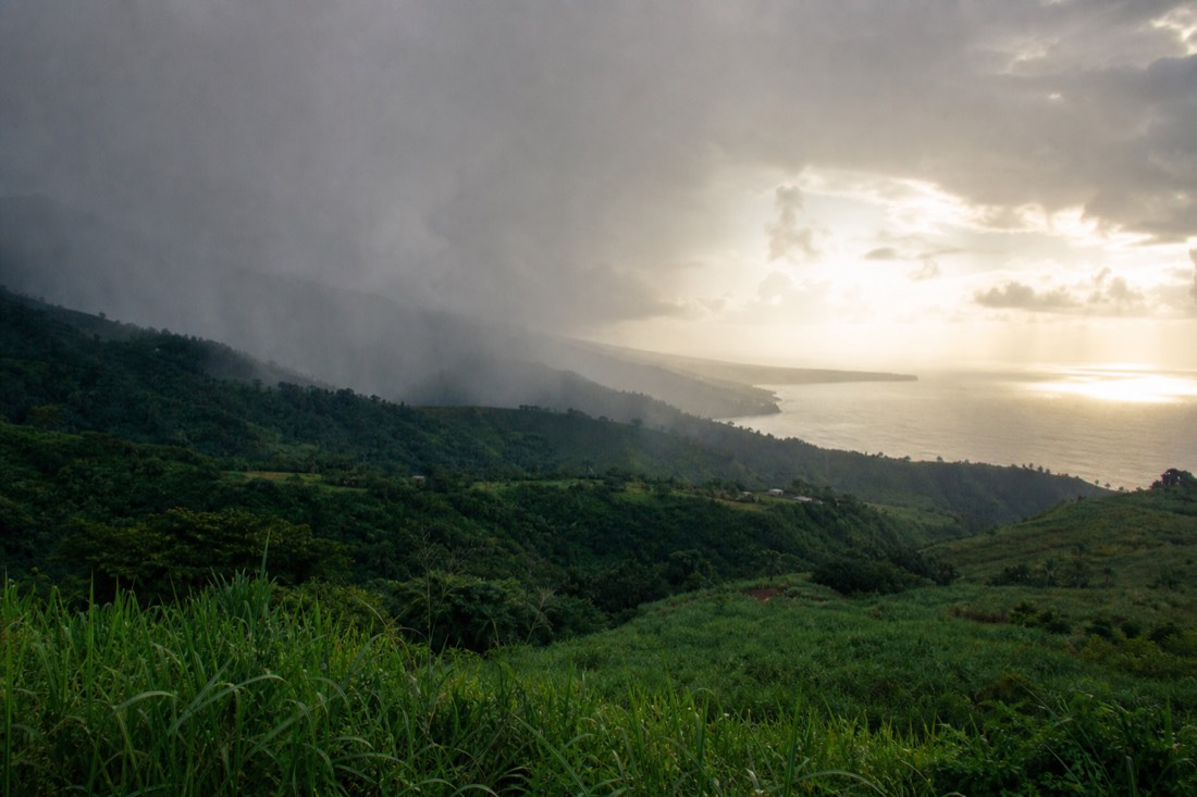 A grey curtain of rain sliding down the Hamakua coastline over dark green jungle and sugar-cane hillsides, sunlight breaking through behind it over the ocean