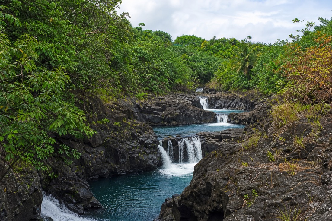 Chain of large round river pools at Boiling Pots on the Wailuku River, water churning between dark basalt ledges under a bright cloudy sky