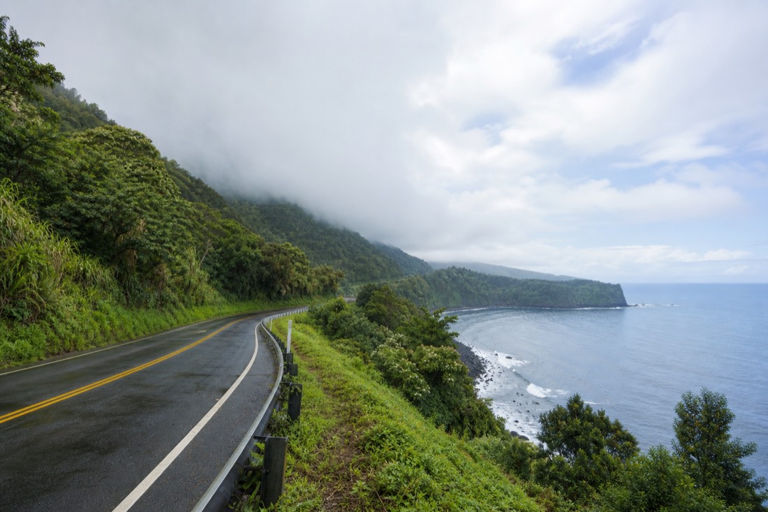 Highway 19 curving north along the Hamakua Coast above the ocean, sugarcane-era greenery and low cloud over the slope, wet asphalt after rain