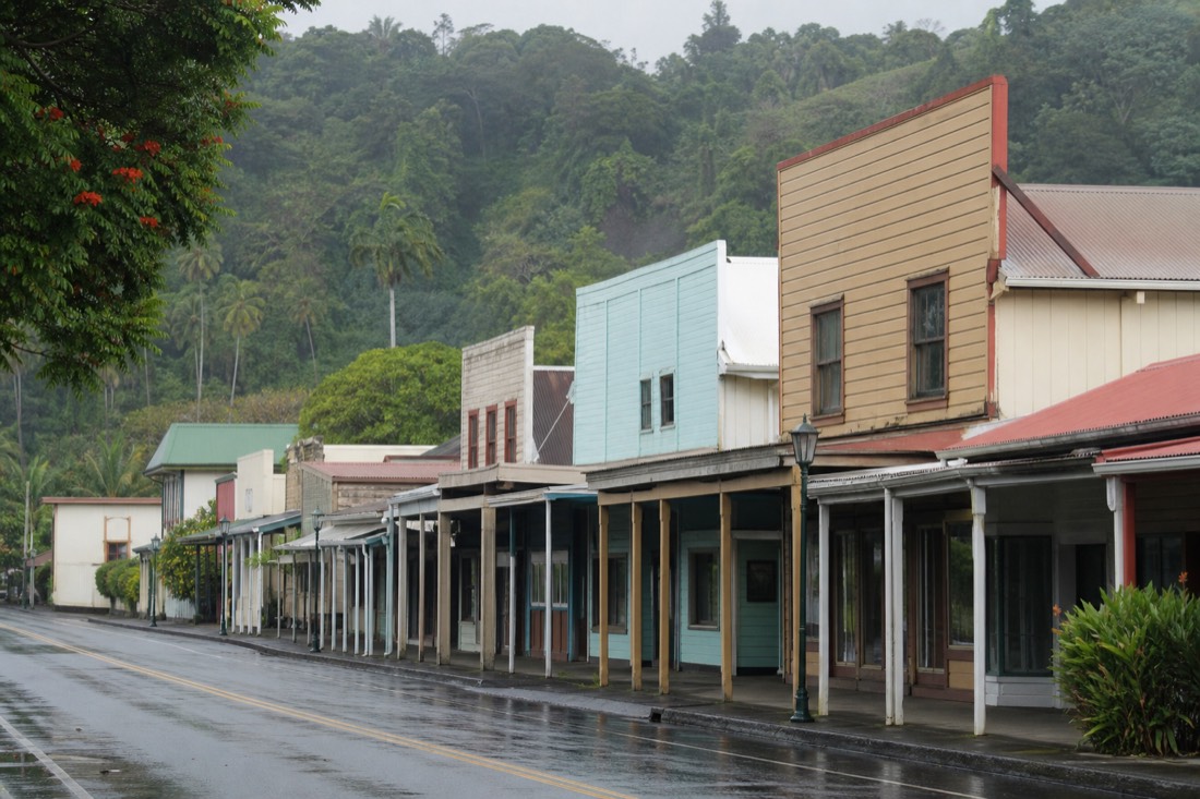 A row of low early-1900s wooden storefronts with deep shaded awnings along a quiet main street in a small tropical town, painted timber facades in faded pastels, green hills rising behind the rooftops, wet pavement after a passing shower. Downtown Hilo, Big Island of Hawaii. No people in focus, no readable signage, no logos.