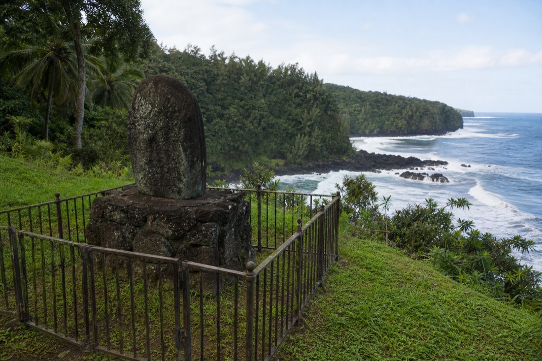 Granite memorial stone on a lava pedestal inside an iron railing on a grassy knoll above Laupahoehoe Bay with surf breaking on black rock below